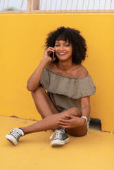 Smiling young female with curly hair wearing stylish outfit sitting on yellow surface and speaking on mobile phone - ADSF47235