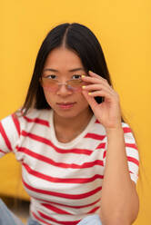 Positive Asian young female in striped tshirt and jeans hile looking at camera with serious while lean on against yellow background - ADSF47236