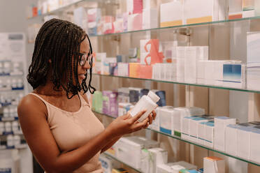 Side view of African American young woman with dreadlocks reading ingredients on medicine bottle label while standing by different products on shelf at drugstore - ADSF47238