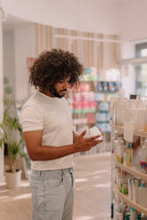 Side view of African American young man with afro hairstyle reading ingredients on medicine bottle label while standing by different products on shelf at drugstore - ADSF47239