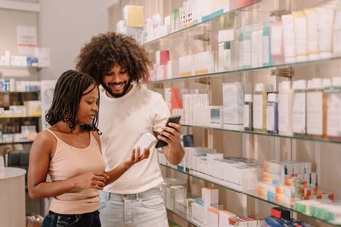 Junger schwarzer Freund mit Afro-Haar und Freundin mit Dreadlocks halten ein Medikament in der Hand und lesen Informationen auf ihrem Handy, während sie in einer Apotheke einkaufen - ADSF47240