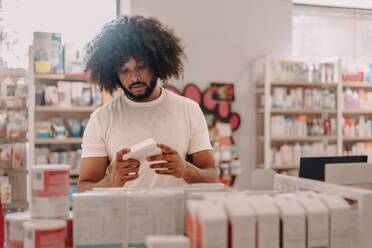African american man with afro hairstyle reading information on prescribed medicine package while standing against shelves with various drugs at pharmacy store - ADSF47242