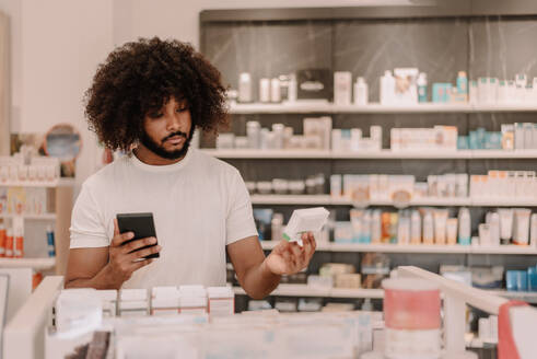 Afroamerikanischer Mann mit Afrofrisur, der ein Smartphone hält und Informationen auf einer Medikamentenpackung liest, während er vor einem Regal mit verschiedenen Medikamenten in einem Apothekengeschäft steht - ADSF47243