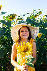 Portrait of cute smiling preteen girl wearing hat and holding beautiful sunflower while standing in farm during summer at weekend - ADSF47254