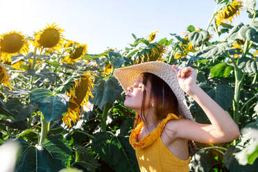 Side view of Caucasian cute preteen girl with eyes closed wearing hat and smelling sunflower growing in farm during summer at weekend - ADSF47255
