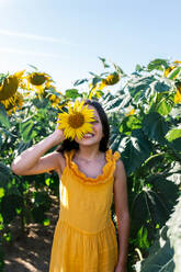 Smiling Caucasian preteen girl dressed in yellow casuals hiding face with beautiful sunflower while standing amidst plants in farm - ADSF47256