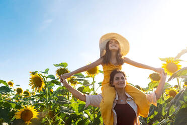 Caucasian mid adult mother carrying smiling adorable daughter on shoulders while standing in sunflower field against clear sky during summer - ADSF47258