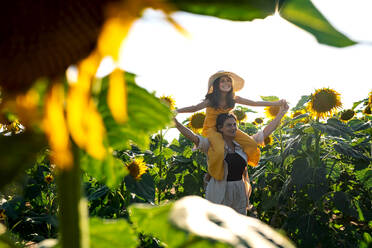 Caucasian mid adult mother carrying smiling adorable daughter on shoulders while standing in sunflower field against clear sky during summer - ADSF47259