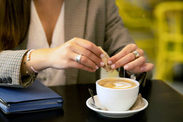 Crop anonymous young female in smart formal clothes sitting at table with diary book while adding brown sugar in creamy coffee in cafe with blur background - ADSF47262