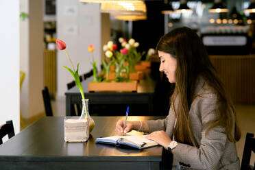 Side view of smiling young female with long hair looking down while sitting at table with notebook and taking notes with pen in cozy blurred cafe - ADSF47264