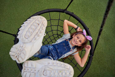 Top view of joyful kid in overall looking at camera while lying on web swing with raised legs on playground in summer - ADSF47265