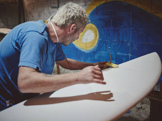 Side view of concentrated bearded male carpenter measuring thickness of edges of surfboard with caliper in workshop - ADSF47269