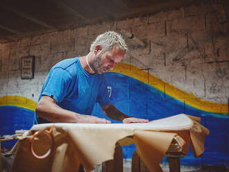 Side view of bearded blond adult male master wrapping handmade surfboard in fabric while decorating board in workshop - ADSF47270