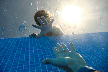 Low angle of son leaning on edge of tiled swimming pool in resort and reaching out hands with crop father on weekend summer day - ADSF47272