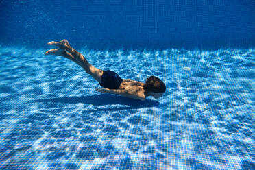 Full length of carefree barefoot boy in swimwear swimming at bottom of tiled pool during summer vacation - ADSF47274