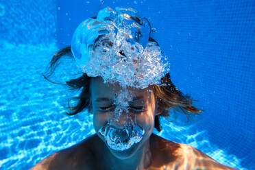 Underwater view of happy child having fun and blowing bubbles while diving into blue swimming pool and smiling - ADSF47275