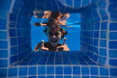 Underwater excited child having fun while swimming in blue pool and looking at camera on weekend summer day - ADSF47277