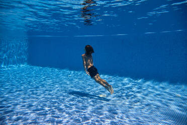 Back view of unrecognizable child in swimsuit diving in blue swimming pool in resort on sunny day during summer vacation - ADSF47278