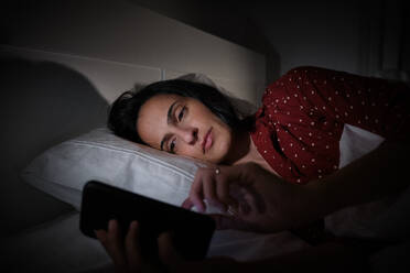 Focused Hispanic female in sleepwear text messaging on modern cellphone while lying on comfortable bed late at night in bedroom - ADSF47280
