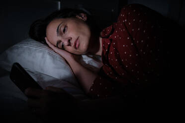 Serious Hispanic female surfing internet on modern cellphone on bed late at night while suffering from insomnia in dark bedroom - ADSF47281