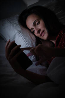 Hispanic female surfing internet on modern cellphone while lying on comfortable white bed in dark bedroom at night time - ADSF47282