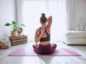 Back view of anonymous female practicing Gomukhasana posture while sitting on mat during yoga training in light living room with couch - ADSF47291