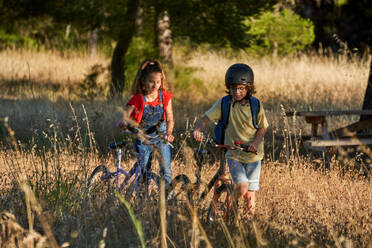 Full length of girl and boy in protective helmet bonding while walking through dry grass with bikes near wooden picnic table placed in recreation area in countryside - ADSF47300