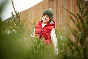 Low angle of delighted little boy in stylish warm clothes and hat smiling and looking at camera while sanding amidst coniferous trees in outdoor local market - ADSF47305