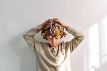 Curly haired sad preschool boy with drooping head covered with sparkles and confetti on white background - ADSF47307