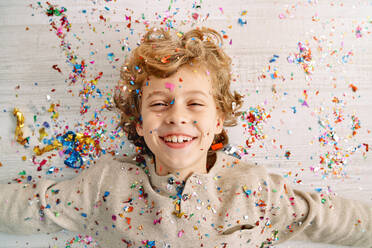 Cheerful preteen boy with blond curly hair lying on light laminated floor covered with multicolored confetti and spangles - ADSF47308