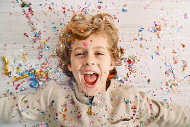 Top view of delighted preteen little boy with curly blond hair lying on laminated floor and screaming wow while enjoying falling confetti and sparkles in party - ADSF47310