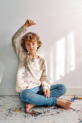 Full body of preteen barefoot boy wearing casual jumper and jeans sitting on floor with crossed legs and throwing colorful confetti - ADSF47311