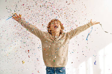 Excited preteen boy with curly hair rejoicing over surprise and screaming happily while standing under falling shiny spangles and confetti - ADSF47312