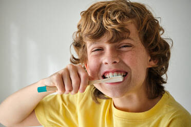 Child with brown wavy hair in yellow wear brushing teeth with toothbrush while looking at camera - ADSF47323