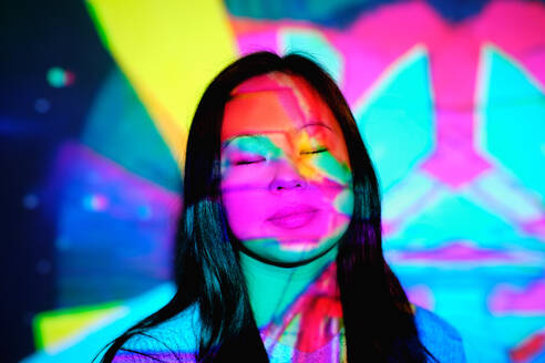 Charming Asian female with long hair standing with closed eyes under bright glowing neon lights on face in stylish studio - ADSF47326