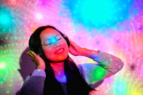Positive Asian female with closed eyes listening to music in wireless headphones while standing near wall with glowing bright neon lights in studio - ADSF47327