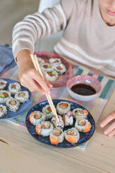 From above of unrecognizable hungry kid eating sushi rolls with soy sauce using wooden chopsticks in dining room at home - ADSF47331