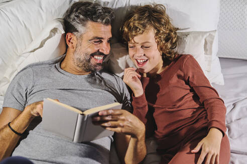 From above of happy father and son in pajamas having fun while reading interesting book and lying on soft bed together at home - ADSF47334