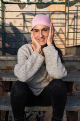 Charming adult Hispanic female in pink headscarf symbol of international breast cancer campaign sitting on grunge while touching face and looking at camera - ADSF47335