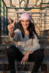 Smiling Hispanic female in pink headscarf of breast cancer awareness campaign sitting on metal stairway and showing two fingers gesture while looking at camera - ADSF47336