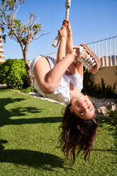 Full body of smiling preteen girl in casual clothes hanging upside down from swing rope and looking at camera in park on sunny day - ADSF47339