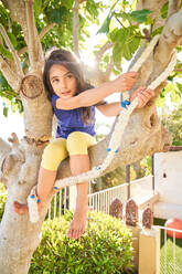 Full body of positive girl in casual clothes playing on tree branch and touching rope swing while looking away in backyard on weekend summer day - ADSF47340