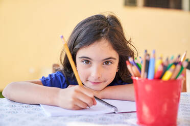 Inspired little child from primary school using pencil for writing on white paper and sitting at desk while looking at camera - ADSF47342