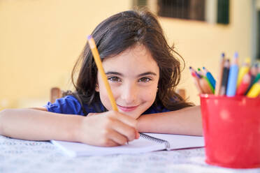 Creative cute schoolgirl taking notes on paper with pencil in copybook while spending time at home and looking at camera - ADSF47345