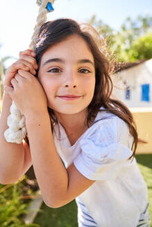 Smiling cute cheerful girl in white casual clothes looking at camera and standing next to rope outside at daylight - ADSF47348