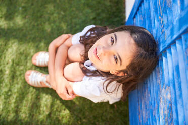 Top view of happy adorable girl looking at camera and sitting on green grass in park during summer weekend - ADSF47349