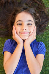 Top view of positive child with long hair touching face and looking at camera while lying on green lawn on weekend summer day - ADSF47351