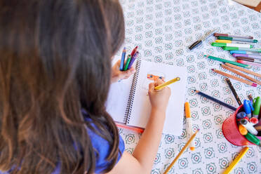 From above back view of creative child sitting at table and drawing picture in notepad with colorful pencils at daytime - ADSF47352