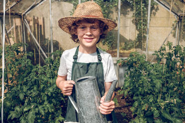 Happy smiling preteen boy in apron and wicker hat standing in greenhouse with watering can and looking at camera - ADSF47376