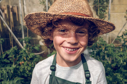 Happy smiling preteen boy in straw hat and apron in hothouse and looking at camera in sunlight - ADSF47377